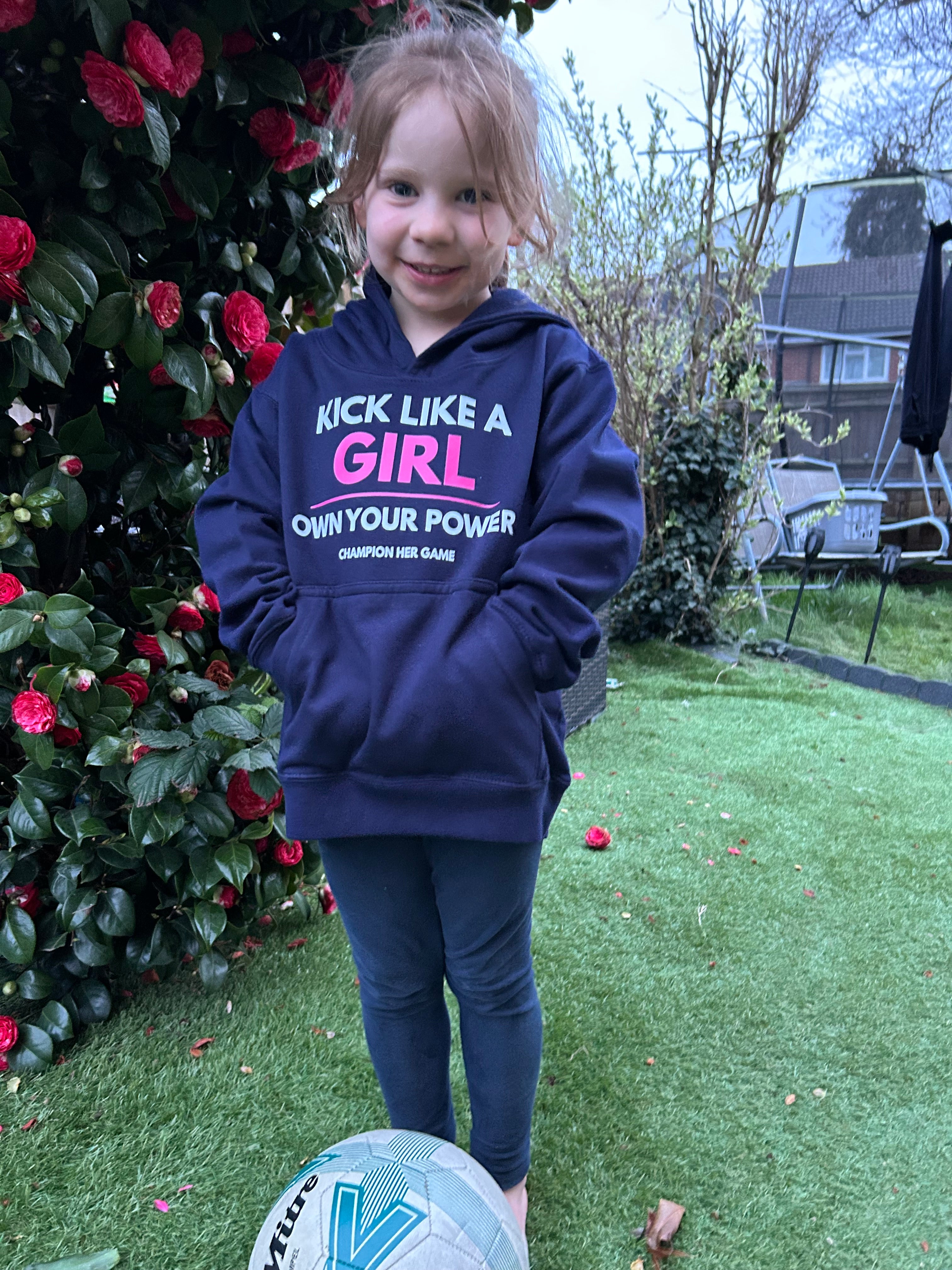 Child wearing a blue hoodie with kick like a girl, standing outdoors with a garden background.