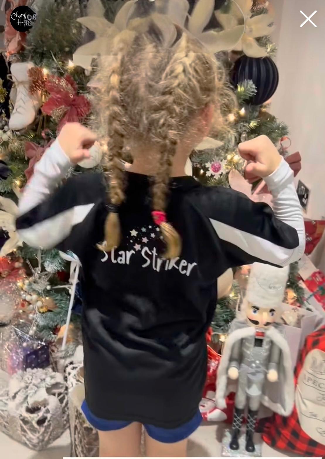 Child wearing a black 'Star Striker' shirt with arms flexed in front of a decorated Christmas tree.