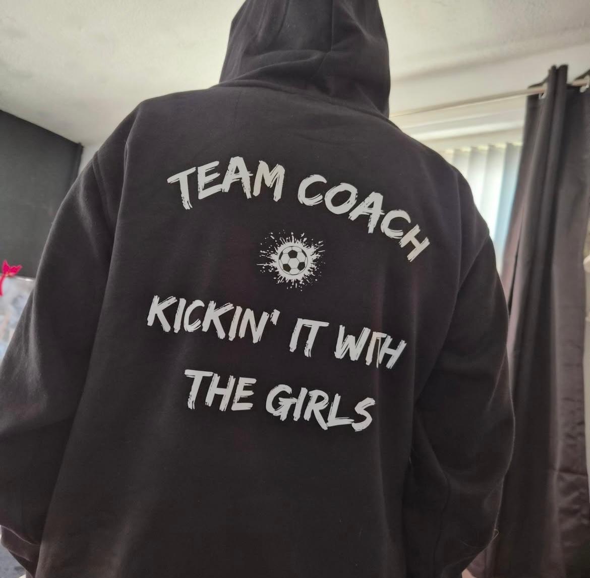 Black hoodie with text on the back worn by a person, indoors.