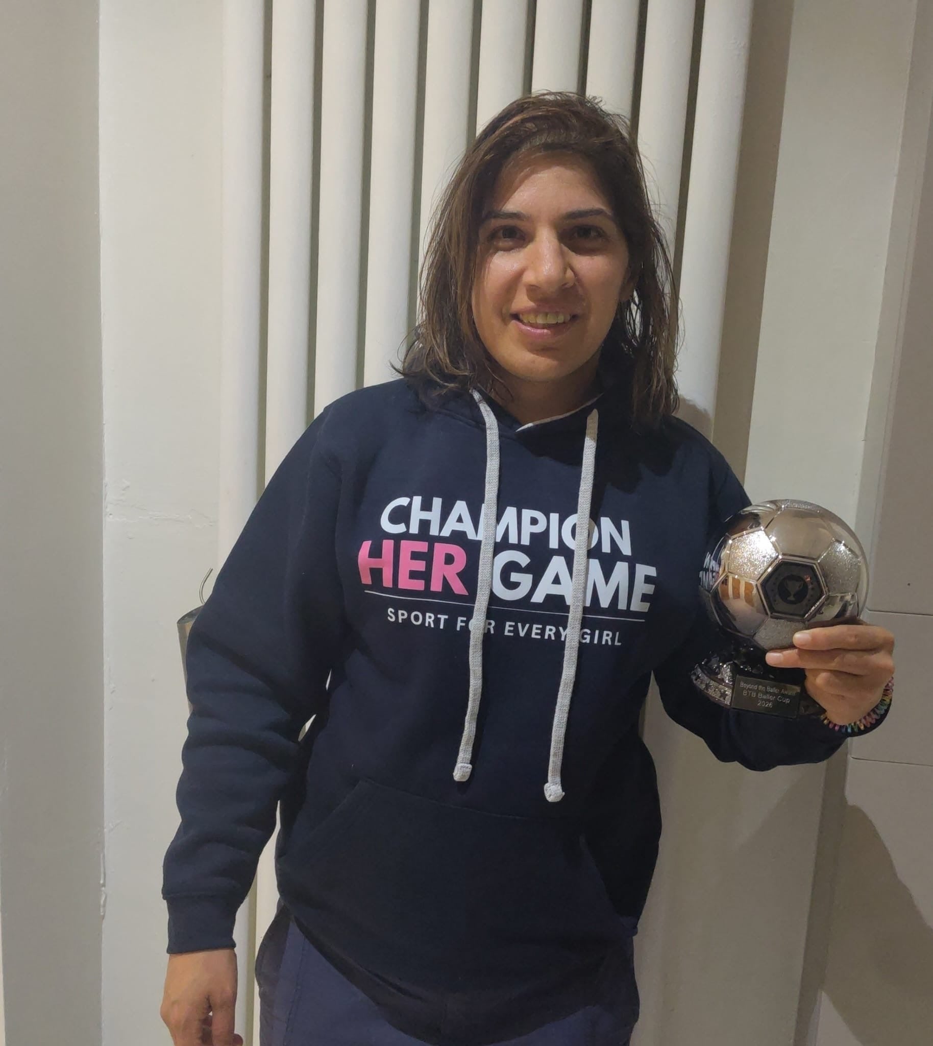 Women wearing a hoodie with 'Champion Her Game' text, holding a soccer ball indoors.
